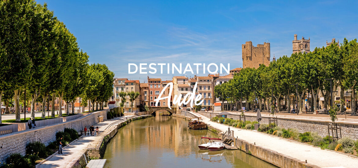 Vue pittoresque du canal bordé d'arbres et de bâtiments historiques dans l'Aude, avec une petite embarcation amarrée au premier plan. Le texte "Destination Aude" est superposé sur l'image.