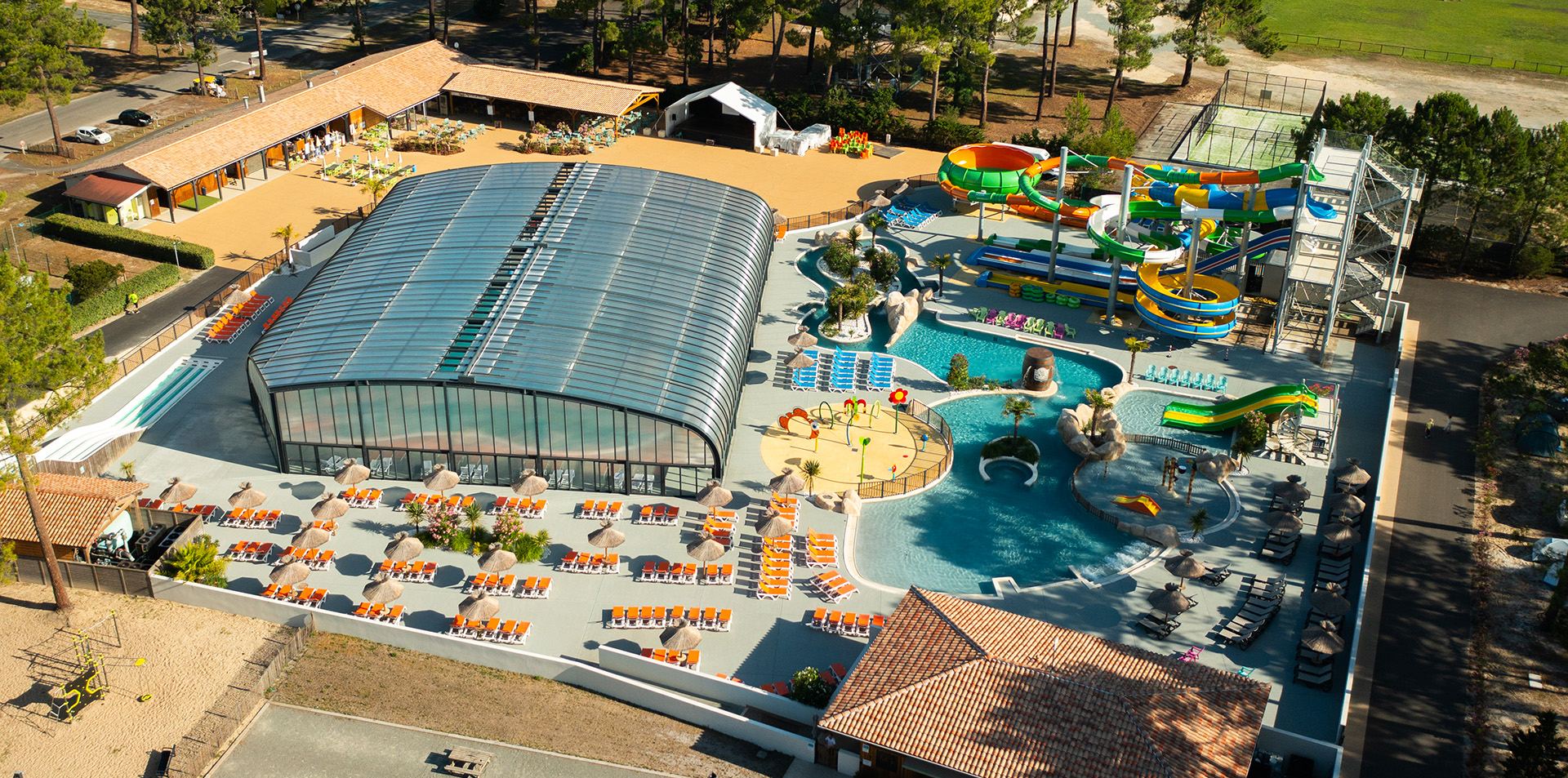 Vue aérienne d'un complexe aquatique avec piscines extérieures, toboggans et espace détente avec des transats sous des parasols.