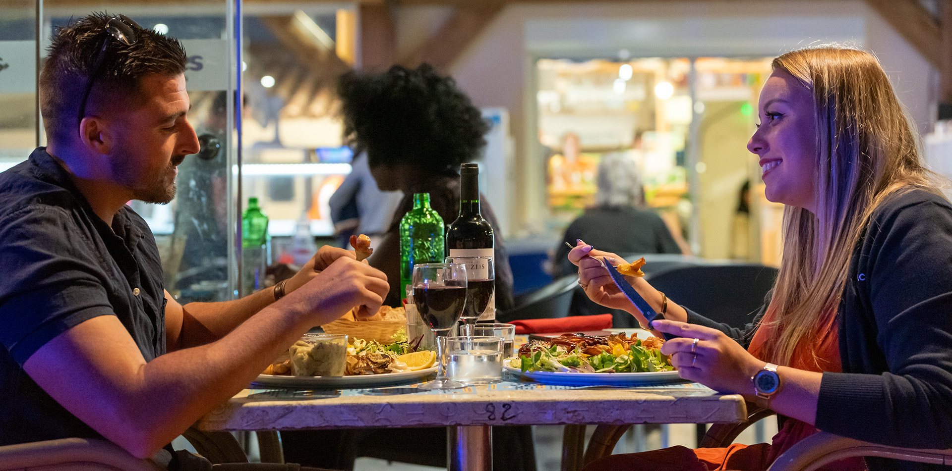 Un homme et une femme souriants en train de dîner dans un restaurant au Camping Campéole La Côte des Roses, avec des assiettes de nourriture et une bouteille de vin sur la table.