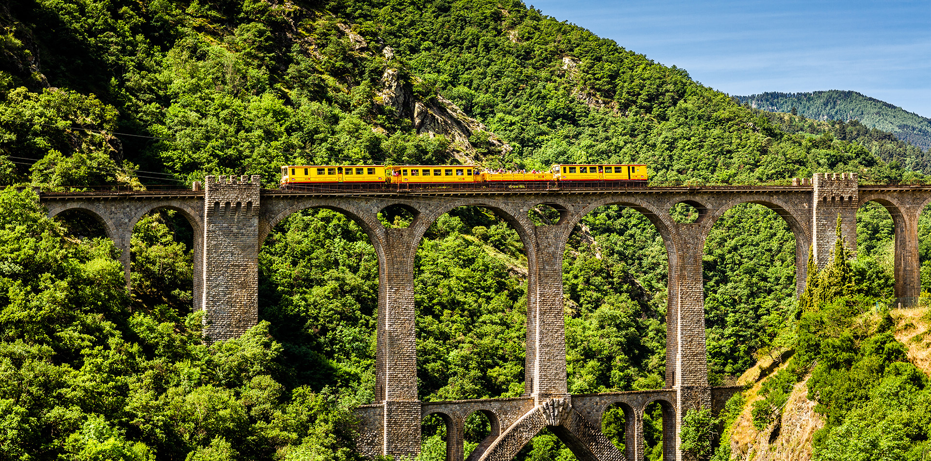 Le train jaune des Pyrénées-Orientales traversant un grand viaduc en pierre au milieu d'un paysage boisé.