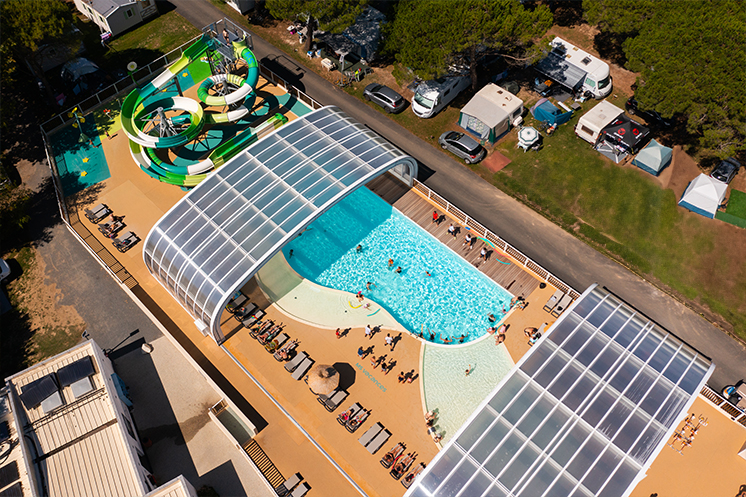 Vue aérienne du parc aquatique du Camping Campéole Pontaillac Plage 4* avec deux grands toboggans verts, une piscine couverte par une structure en demi-cercle transparente, et des visiteurs se prélassant sur des chaises longues.