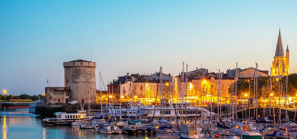 Panorama du crépuscule sur le port de La Rochelle avec les Tours de la Chaîne et Saint-Nicolas, des bateaux amarrés et l'illumination des façades et des monuments historiques en arrière-plan.