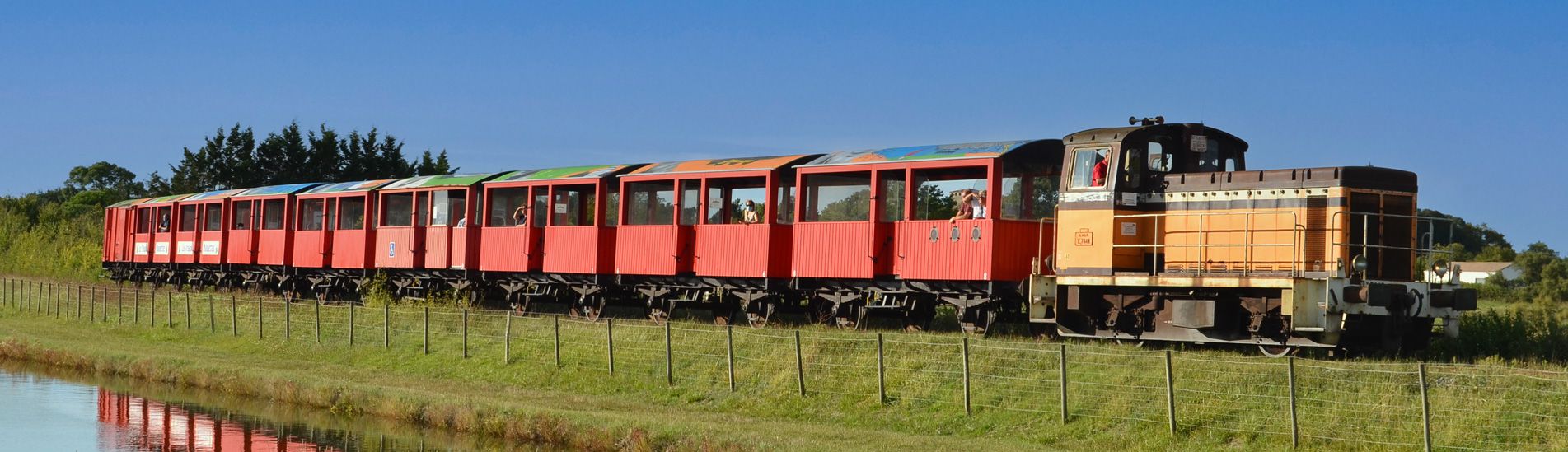 Le Train des Mouettes, train touristique à vapeur avec des wagons rouges traversant une zone rurale ensoleillée avec un plan d'eau au premier plan.