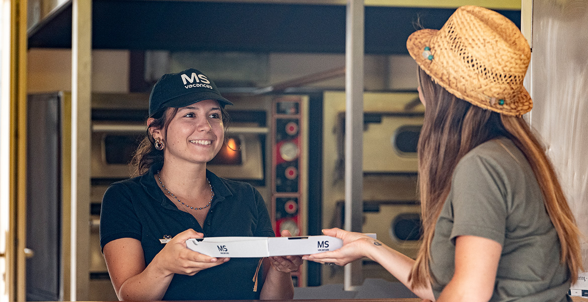 Employée souriante de MS Vacances remettant une pizza à une cliente ayant payer avec son bracelet cashless.