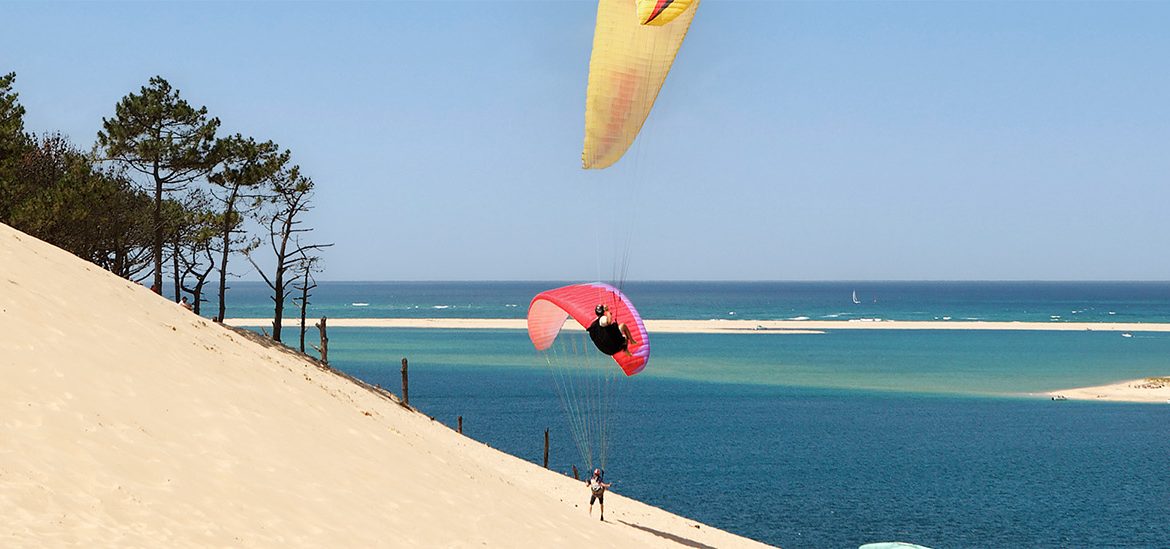Plusieurs parapentes colorés survolent la dune du Pilat, avec une vue sur l'océan Atlantique et une lagune en contrebas, entourée de pins.