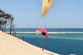 Plusieurs parapentes colorés survolent la dune du Pilat, avec une vue sur l'océan Atlantique et une lagune en contrebas, entourée de pins.