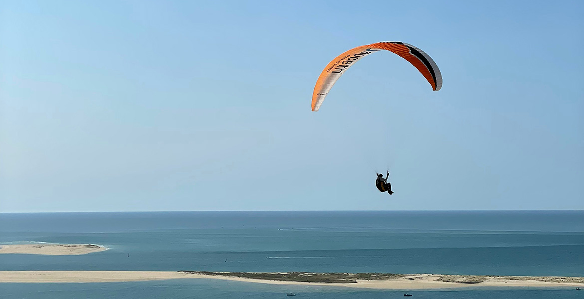Un parapentiste sur la dune du Pilat plane au-dessus des bancs de sable et des eaux calmes du bassin d’Arcachon, sous un ciel dégagé.