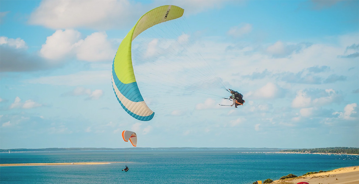 Des parapentistes volent au-dessus de la dune du Pilat sous un ciel partiellement nuageux, profitant d’une vue panoramique sur le bassin d’Arcachon.