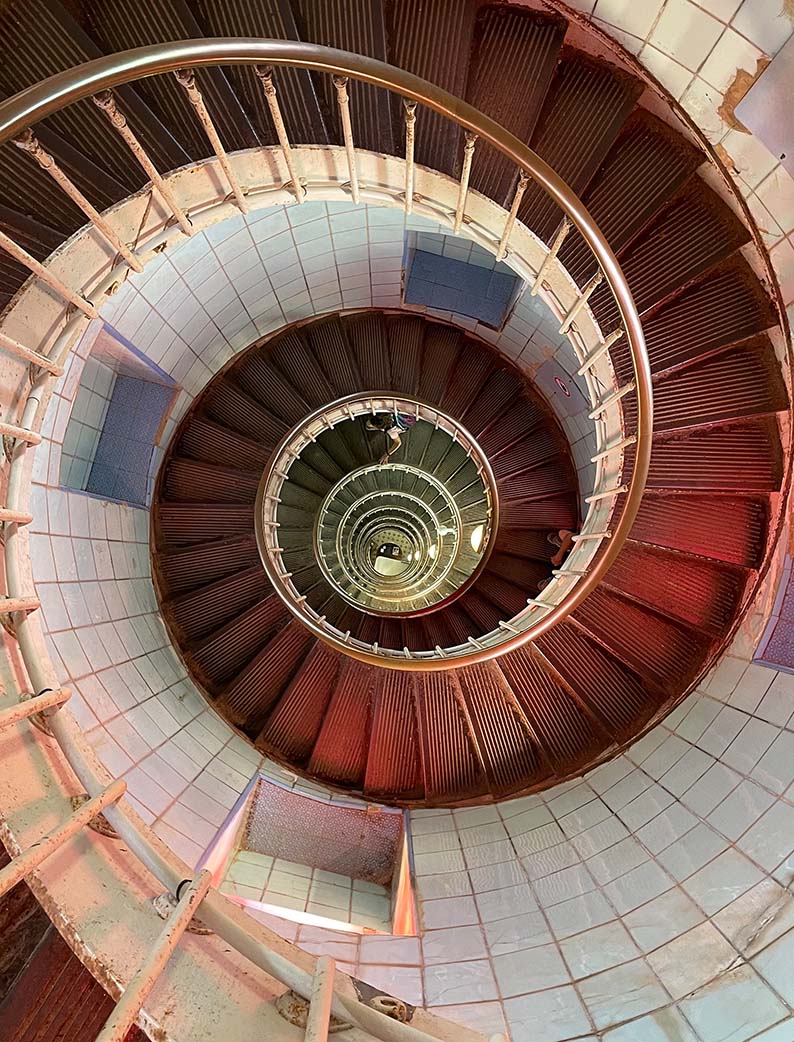Escalier en colimaçon de l'intérieur du phare de la Coubre, vu en plongée depuis le haut du phare, avec une rampe métallique et des marches rougeâtres.