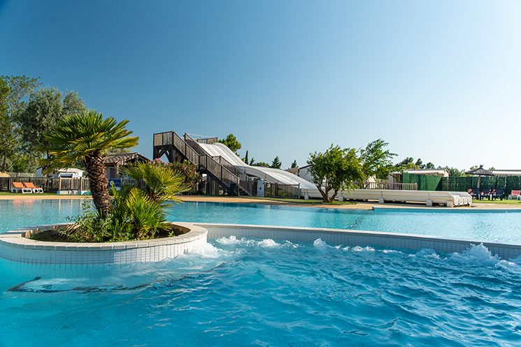 Piscine extérieure avec bains à remous, palmier sur îlot central, toboggan aquatique en arrière-plan avec ciel bleu dégagé.